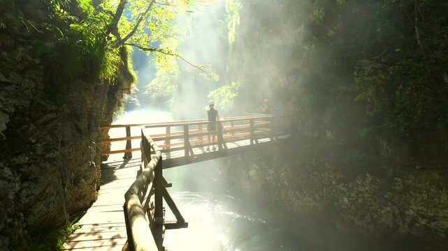 Sunbeams illuminating mist rising from radovna river in vintgar gorge near bled, with tourist walking on wooden footbridge