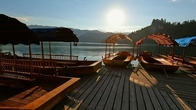 Wooden tourist boats moored at dock on lake bled in slovenia with bled island and iconic church in background and sun shining through canopy