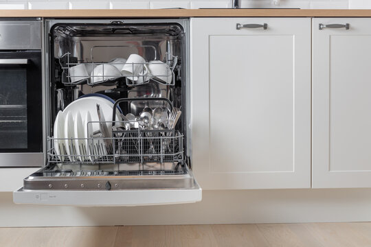 Built-in dishwasher full of clean dishes with its door open, white utensil. Modern technologies in the home kitchen with minimalistic cabinetry. Simplifying daily housework.