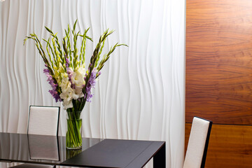 A bouquet of white and purple lilac gladiolus flowers in a glass vase on a glass table with reflections, opposite a white textured wall.