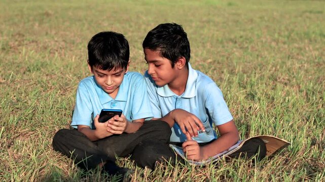 Two elementary age cheerful children of Indian ethnicity wearing school uniform sitting outdoor in nature holding book and using mobile phone together video portrait close up.