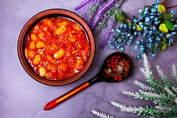 Bean soup, beans in tomato in a clay plate, wooden spoon, berries and herbs on a purple background