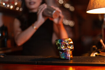 Skull glass stands on bar counter. Girl bartender makes cocktail with shaker in bokeh