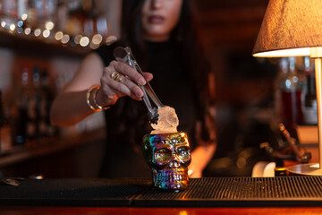 Girl bartender makes alcoholic cocktail and puts ice cube in glass skull on bar counter
