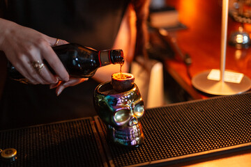 Girl bartender prepares alcoholic cocktail pours liqueur into glass skull with passion fruit on bar counter