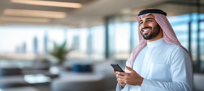 Smiling Middle Eastern businessman in traditional clothing using his phone in a modern office.