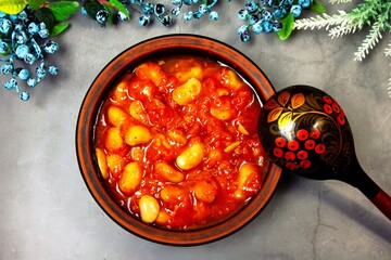 Bean soup, beans in tomato in a clay plate, wooden spoon, berries and herbs on a purple background