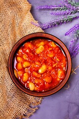 Bean soup, beans in tomato in a clay plate, burlap, berries and herbs on a purple background