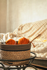 Vertical image. Orange knitting yarns and needles in the basket against sofa. Cozy mood. Home leisure