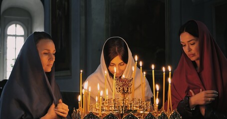 Three women pray in serene candlelit church, embodying devotion and spirituality. Holy atmosphere reflects Orthodox traditions and inner peace, illuminated by soft glow of candles and icons.