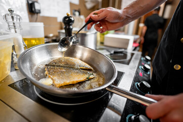 A chef expertly cooks fish in a stainless steel pan, using a spoon to baste it with sizzling oil....