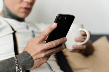 Handsome young man with tattoos relaxes on a couch, sipping coffee and using his smartphone.