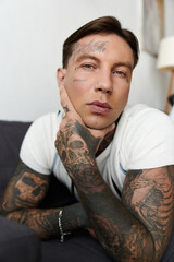 Handsome young man with tattoos sits calmly, resting his chin on his hand in a relaxed atmosphere.