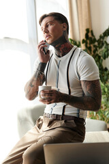 Handsome young man with tattoos engages on a phone call while seated and holding a cup.