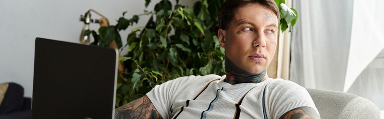 Handsome young man with tattoos focused on laptop, sitting comfortably indoors with plants.