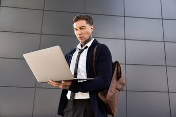 Handsome man with tattoos is focused on his laptop while standing in a contemporary setting.