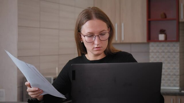 Businesswoman wearing glasses is sitting at her laptop computer in her home office and comparing information on paper documents with data on her screen