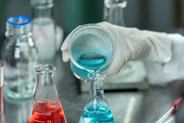 A gloved hand carefully pouring a blue liquid from a beaker into an Erlenmeyer flask