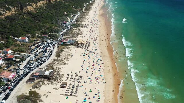 Aerial drone footage showing packed beach at Praia de Fonte da Telha in Costa de Caparica, Portugal. People sunbathe and swim while the adjacent parking lot is filled with cars. Beach season 