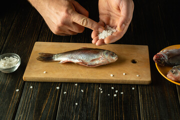 Skilled hands sprinkle coarse salt over a fresh fish on a wooden cutting board, setting the stage for a flavorful dish. Nearby, additional fish await their turn in the kitchen