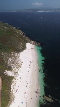 Ascent over Melide Beach on Ons Island on summer morning: turquoise waters, white sand, tourists relaxing, lush dunes, rugged rocks, a dirt path, and view of Magor Beach across the Pontevedra estuary.