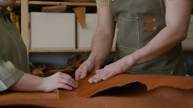 Unrecognizable male master craftsman in apron and his female apprentice touching piece of genuine leather on table at workshop