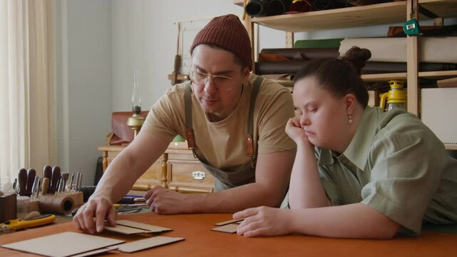 Master craftsman and his young female apprentice with Down syndrome choosing pattern to work with in leather workshop
