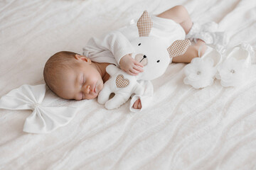 A baby is sleeping on a bed with a stuffed rabbit next to it. The baby is wearing a white outfit and has a bow in its hair
