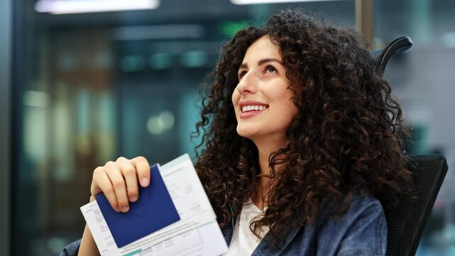 Excited woman with curly hair sits in an airport lounge and dreams of her next adventure holding her passport and ticket.