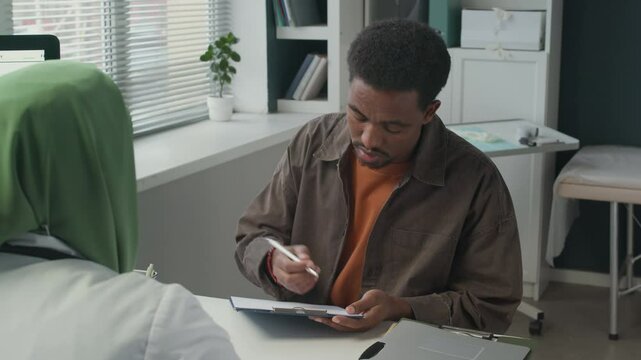 Medium shot of young African American male patient sitting at table with Muslim female doctor and filling out medical form in modern clinic in daytime