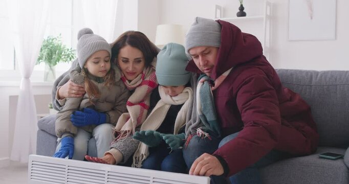 Parents, small children freezing at home wearing winter warm clothes sitting close near radiator heater, badly shivering with strong winter cold at home, low temperature problem, dissatisfied tenants