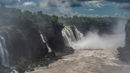 Obraz premium A stormy river in the gorge. Turbulent flows are foaming. A powerful waterfall collapses from a cliff into the riverbed. Splashes, fog. Lush tropical vegetation on the rocks. Clouds in the blue sky.