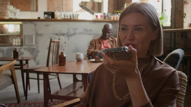 Tilt up of Caucasian modern woman sitting comfortable at cozy cafe table with cappuccino and dessert, recording and sending audio message via smartphone