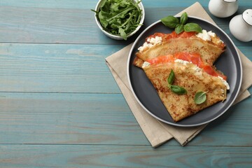 Delicious crepes with salmon, cream cheese, basil and arugula on light blue wooden table, flat lay....