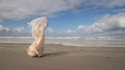 art portrait of woman in long white dress near blue ocean on the beach holding thin fabric on windy day