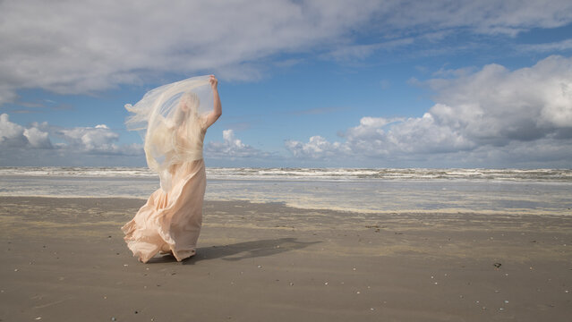 art portrait of woman in long white dress near blue ocean on the beach holding thin fabric on windy day