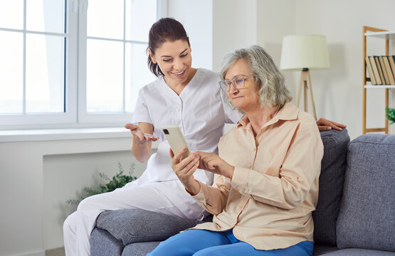 Caregiver assists an elderly woman with mobile phone and online technology at a nursing home. Nurse offers support with internet usage, ensuring the patient access to modern communication tools.