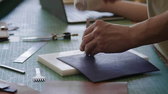 Close-up of unrecognizable craftsman using mallet and leather-stitching chisel to work with dark blue leather on cluttered workbench in studio