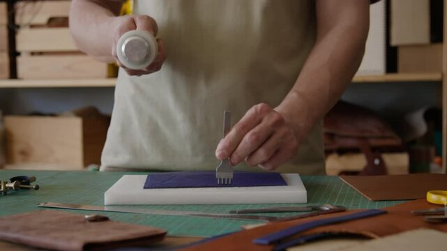 Unrecognizable craftsman using mallet to strike stitching chisel against leather on white cutting board in workshop
