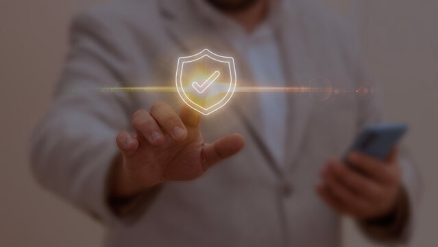Businessman interacting with a glowing shield icon featuring a checkmark, symbolizing data protection, security, and trust in digital environments and professional technology systems.