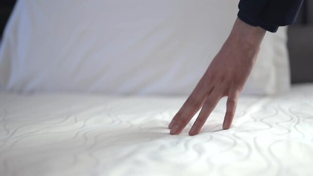 Unknown hand carefully presses down on a plush, white mattress, showcasing its softness and comfort. Closeup of unrecognized person hand arranging bed in hostel suit.