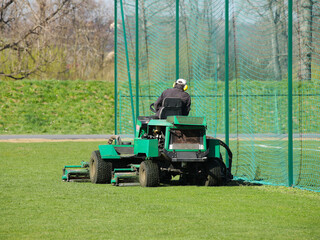 Worker on a large green lawn mower mows the grass on the football field. Landscape design and maintenance of green areas of the sports complex. Making grass stadium before the match. Human labor