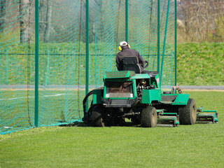 Worker on a large green lawn mower mows the grass on the football field. Landscape design and maintenance of green areas of the sports complex. Making grass stadium before the match. Human labor