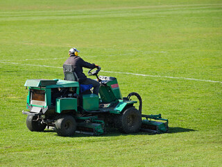 Worker on a large green lawn mower mows the grass on the football field. Landscape design and...