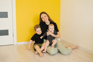 Young happy mother and her two children are sitting on the floor in front of a yellow wall. The scene conveys a warm and loving family moment