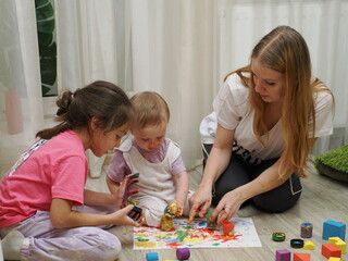 Mom plays with children, funny baby shows his palms painted with paint. Creative art classes. Laughter. Happy children and mom finger painting on paper at home. Bright colors, gouache and watercolors