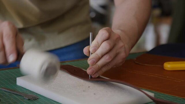 Close-up shot of unrecognizable craftsman meticulously punching holes in leather belt using mallet and chisel in workshop