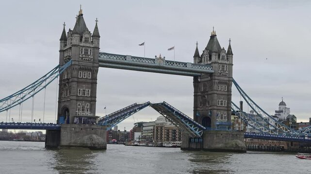 Tower Bridge's bascules are raised for a boat, set against an overcast sky. Gothic towers and cityscape blend tradition and modernity in London.