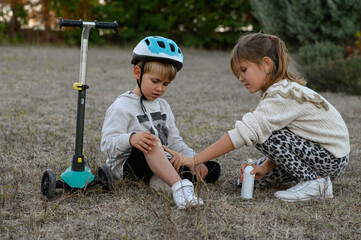 A young boy in a helmet riding a scooter falls and scrapes his knee. A girl approaches him, showing empathy and sprays medicine on his knee to help soothe the injury. 