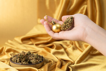 in white young female hand dry bud of medical marijuana and Christmas ball. On festive golden background